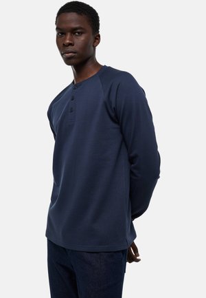 Homme à la peau foncée portant une chemise henley manches longues bleu marine et un pantalon foncé, debout les mains derrière le dos.
