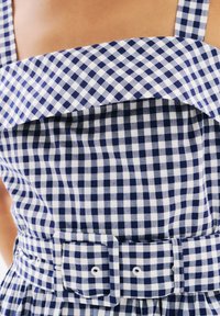 Ruudullinen mekko neliönmallisella pääntiellä, jossa on sinivalkoiset ruudut, vyötäröllä vyö ja liehuva hame. Pintakudos näyttää sileältä ja kevyeltä.