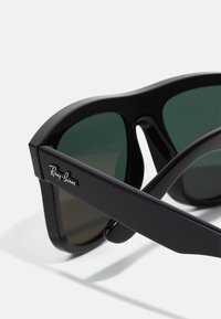 Ochelari de soare negri cu lentile verzi închise, având un cadru gros din plastic. Logo-ul Ray-Ban este vizibil pe braț.