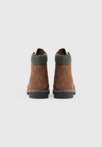 Botas de tobillo de ante marrón con un collar acolchado verde y suelas de goma negras. Presentan un cosido limpio y un logotipo cerca del talón.