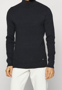 Man som bär en svart ribbad långärmad polotröja och vita byxor, stående framför en enfärgad ljus bakgrund.