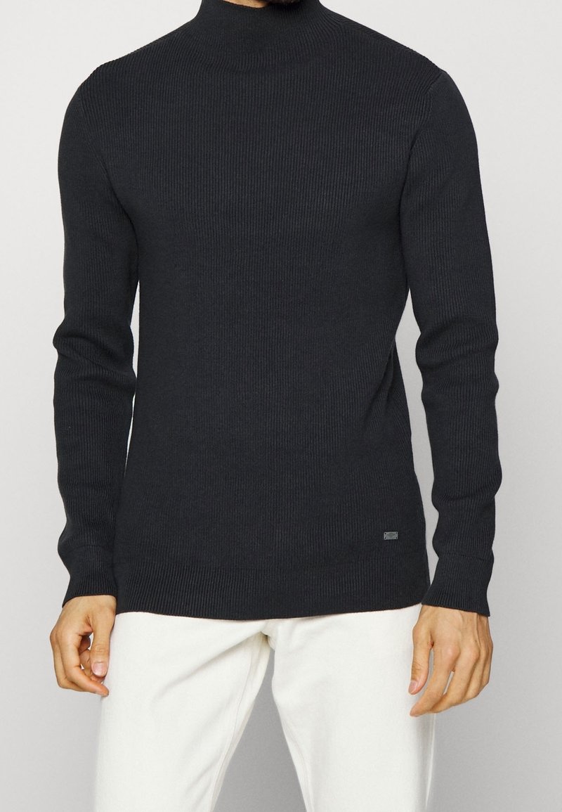 Man som bär en svart ribbad långärmad polotröja och vita byxor, stående framför en enfärgad ljus bakgrund.