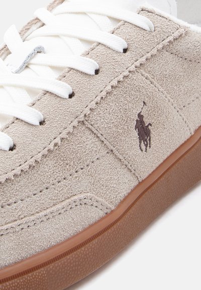 Μπεζ σουέντ αθλητικό παπούτσι με λευκά κορδόνια, καφέ λαστιχένια σόλα και σκούρο κεντημένο λογότυπο πολίστα στη πλαϊνή επιφάνεια.