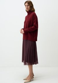 Maglione oversized bordeaux con collo alto abbinato a una gonna midi plissettata con motivo rosso e blu. Scarpe con tacco di colore neutro.