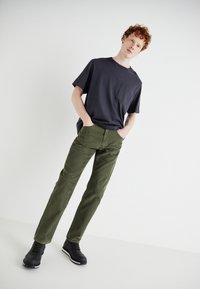 Camiseta gris oscuro con bolsillo, combinada con pantalones verde olivo de corte recto. Zapatos negros con cordones y acentos blancos. Diseño minimalista.