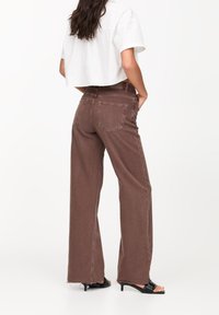 Jean beige à jambes larges avec taille haute et couture contrastante, associé à une chemise blanche courte. Comprend cinq poches et un ourlet légèrement effiloché.