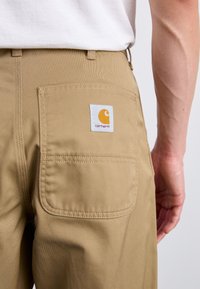 Pantaloni de lucru bej cu un buzunar spate care prezintă o etichetă albă și un logo portocaliu. Textură netedă și material durabil, conceput pentru utilitate.