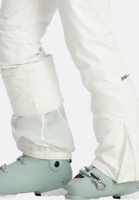 Weiße Skihosen mit verstellbaren Säumen und Reißverschlussdetails, kombiniert mit mintgrünen Skischuhen mit metallischen Schnallen und strukturierten Akzenten.
