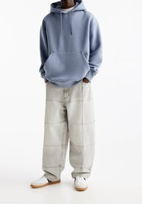 Persona vistiendo una sudadera azul clara, pantalones de patchwork gris claro y sueltos, y zapatillas blancas con suelas de goma, de pie sobre un fondo blanco.