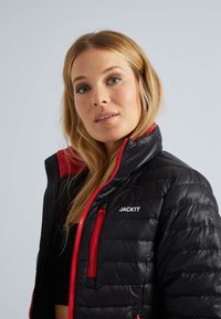 Veste doudoune noire avec des accents et des fermetures éclair rouges, présentant un col montant, un motif matelassé et un logo sur le côté gauche.
