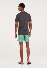 Grå t-shirt med korte ærmer, parret med mintgrønne badebukser med palmeprint. Model iført klipklapper, set fra ryggen.