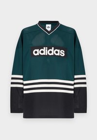 Zöld és fekete Adidas mez V-nyakkal, hosszú ujjakkal, hálós textúrával, a mellkason vastag fehér logóval és csíkos részletekkel.