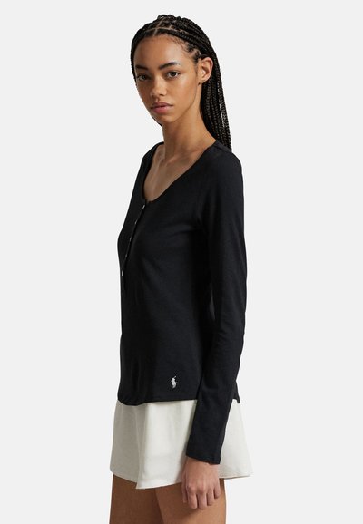 Jeune femme aux cheveux tressés portant un haut henley noir à manches longues et une jupe blanche, debout devant un fond clair uni.