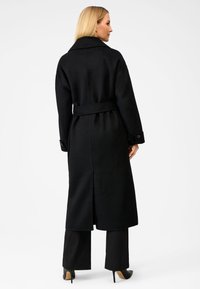 Sort uldfrakke med bælte, oversized krave, lange ærmer og sidelæg. Modellen har sorte bukser og højhælede sko på.