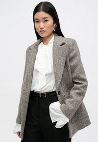 Μπλέιζερ καφέ και μπεζ με σχέδιο καρό, με δομημένους ώμους, κλείσιμο με έναν κουμπί και δύο μπροστινές τσέπες, φορεμένο πάνω από μια λευκή μπλούζα.