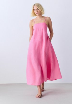 Femme aux cheveux blonds courts portant une robe midi rose sans manches et des sandales marron, debout sur un fond blanc uni.