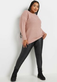 Rosa Strickpullover mit geripptem Muster, kombiniert mit schwarzen Leggings aus Kunstleder und schwarzen Schnürstiefeln. Das Model steht in einer entspannten Pose.