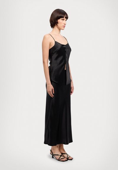 Schwarzes Satin-Cami mit dünnen Trägern, das mit einem Knopfverschluss und einem asymmetrischen Saum versehen ist, kombiniert mit einem passenden langen Rock und Sandalen mit Riemchen.