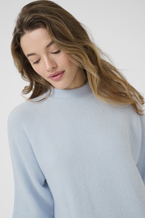 Młoda kobieta o falujących jasnobrązowych włosach, ubrana w jasnoniebieski sweter z dzianiny, patrząca w dół na tle jednolitego szarego tła.