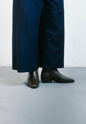 Botas de cuero marrón hasta el tobillo con punta afilada y tacón bajo, combinadas con pantalones oscuros de pierna ancha. Textura suave y ligero brillo.