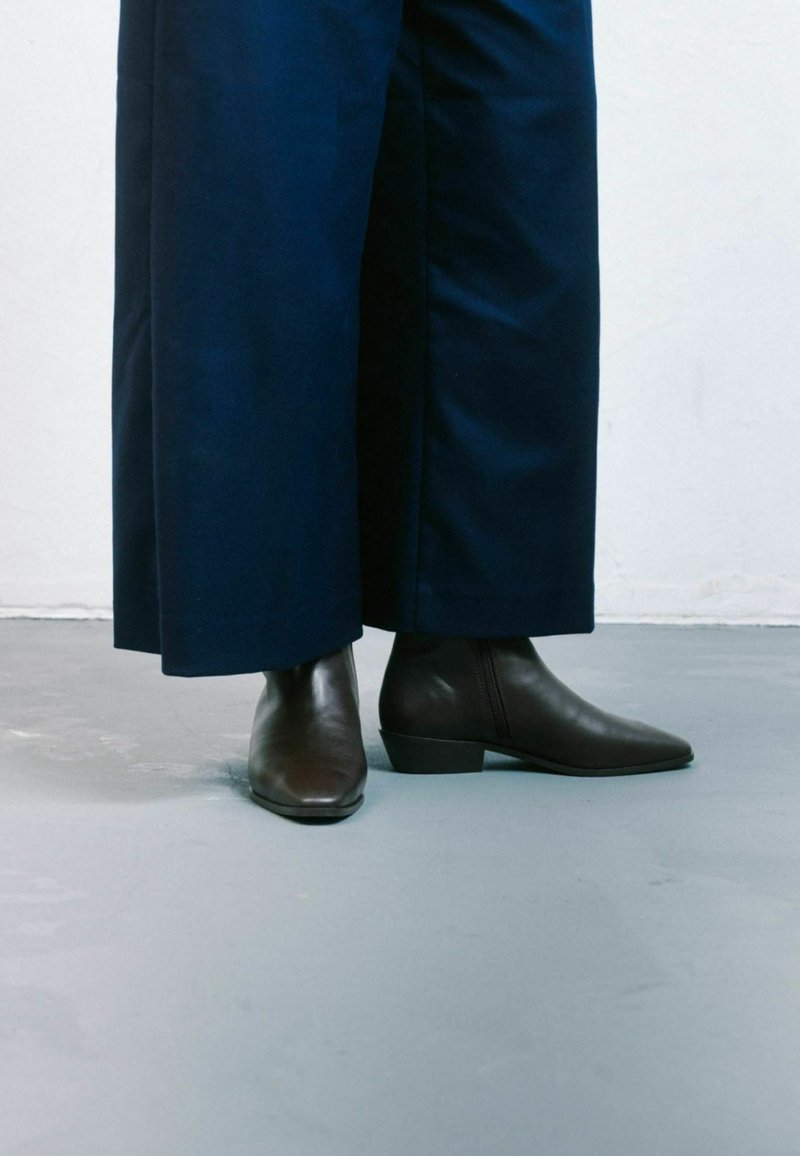 Botas de cuero marrón hasta el tobillo con punta afilada y tacón bajo, combinadas con pantalones oscuros de pierna ancha. Textura suave y ligero brillo.