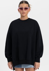 Zwarte oversized sweatshirt met een ronde hals, geribde manchetten en zijsplitten. Draagt een spijkerbroek shorts en gecombineerd met paarse cat-eye zonnebril.