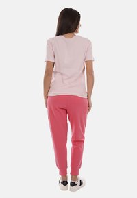 Donna in piedi di spalle, indossa una maglietta rosa chiaro a maniche corte, pantaloni jogger rosa acceso e sneakers bianche su sfondo bianco.