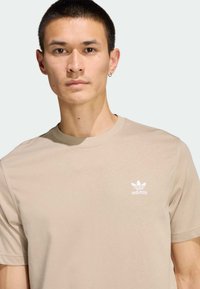 Béžové bavlnené tričko s okrúhlym výstrihom, na ľavej strane na hrudi s malým bielym logom Adidas. Mäkká textúra, krátke rukávy a minimalistický dizajn.