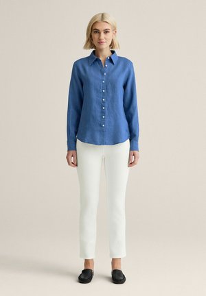 Kvinde stående iført en blå skjorte med lange ærmer og knapper, hvide slim fit bukser, sorte slip-on sko og gyldne hoop øreringe på en beige baggrund.