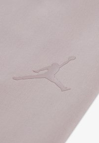 Ανοιχτό ροζ ύφασμα με λεπτό ανάγλυφο λογότυπο σιλουέτας ενός πηδώντας ατόμου, που δείχνει ομαλή υφή και μινιμαλιστικό σχεδιασμό.