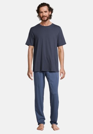 Uomo con la barba che indossa una camicia blu scuro a maniche corte e pantaloni blu, in piedi a piedi nudi davanti a uno sfondo chiaro e uniforme.
