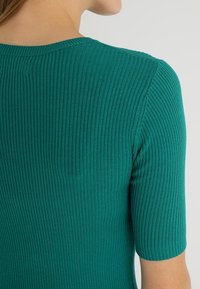 Teal ribbed short-sleeve top featuring a round neckline, fitted silhouette, and textured knit pattern on the fabric.