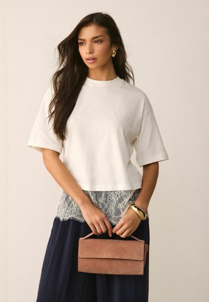 Femeie cu păr lung și închis la culoare, poartă un top alb supradimensionat, fustă bleumarin cu detalii din dantelă, bijuterii aurii și ține în mână o poșetă clutch din piele de căprioară culoarea tabac.