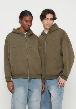 Dva mlada odrasla, oblečena v ujemnih olivno zelenih zadrgnih puloverjih s kapuco in svetlomodrih kavbojkah, stojita drug ob drugem na belem ozadju.