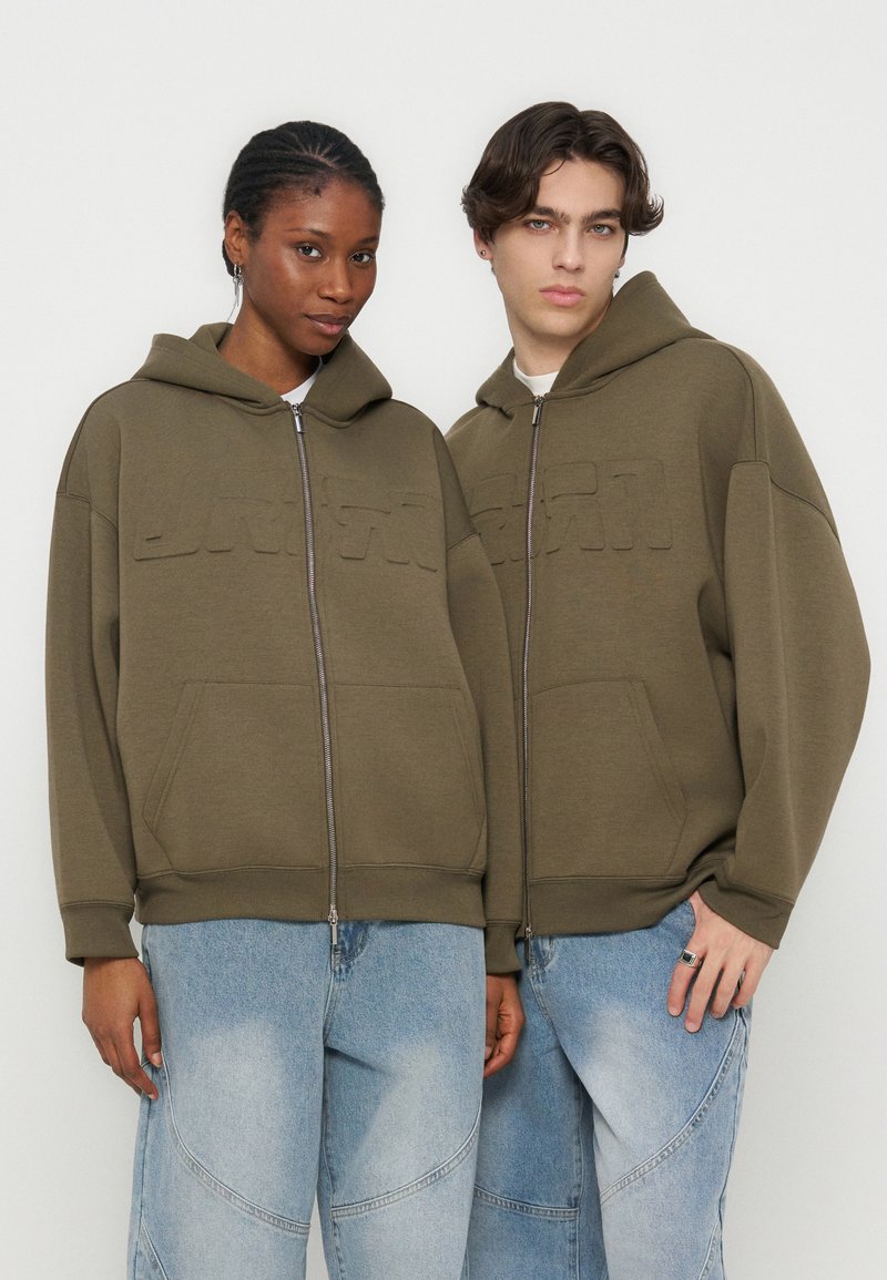 Δύο νεαροί ενήλικες φορούν όμοια ελιάς πράσινα φούτερ με φερμουάρ και ανοιχτά μπλε τζιν, στέκονται δίπλα δίπλα σε λευκό φόντο.