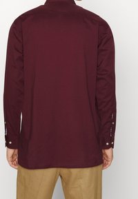 Camisa de manga larga burdeos de algodón, con puños abotonados y dobladillo recto. La vista trasera muestra un diseño simple y limpio.
