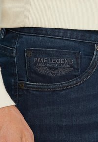 Temno modri jeansi iz denima imajo zadnji žep z vtisnjenim logotipom "PME Legend American Classic" in kovinskimi gumbnimi detajli. Prikaz od blizu.