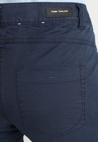 Mörkblå byxor med bakficka med synlig söm och en svart läderapplikation märkt "TOM TAILOR" på midjebandet.