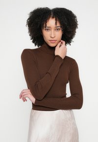 Smeđi rebrasti džemper s rolkom i dugim rukavima, uskog kroja. Model pozira s rukom na vratu, na neutralnoj pozadini.