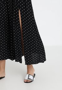 Svart klänning med prickar och en svävande design, med två sidoklyftor. Matchad med justerbara vita sandaler med spänne.