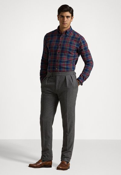 Chemise à carreaux en bleu marine, rouge et vert, associée à un pantalon gris sur mesure. Des chaussures en cuir marron complètent la tenue.