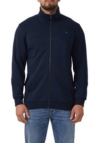 Marineblauwe ritsjack met hoge kraag, ribgebreide manchetten en zoom, met een gladde textuur en een subtiele logo op de borst.