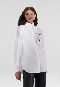 Camicia bianca con colletto a punta, caratterizzata da un design grafico rosso sulla tasca sinistra del petto. Realizzata in tessuto liscio.