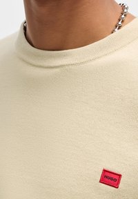 Pull en tricot beige avec col rond, présentant une petite étiquette rouge HUGO sur le côté gauche près de l'ourlet. Texture lisse, design décontracté.