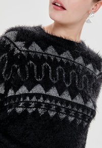 Svart och grå fluffig tröja med geometriska mönster; rund halsringning med lös passform och mjukt, texturerat tyg. Silveraccessoar synlig.