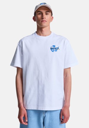 Joven con gorra blanca y camiseta blanca con pequeño gráfico azul en el pecho, combinado con vaqueros azul claro, de pie frente a un fondo liso.