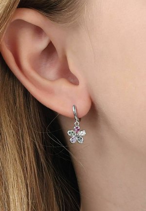 Primer plano de una oreja con un pendiente plateado en forma de flor que presenta piedras preciosas multicolores en el lóbulo, con cabello castaño claro visible.