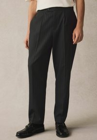Pantaloni neri a righe sottili realizzati in tessuto liscio, con una vestibilità su misura, chiusura con bottone e tasche laterali. Indossati con mocassini neri.