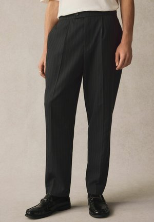 Schwarze Nadelstreifen-Hosen aus glattem Stoff, mit einem taillierten Schnitt, Knopfverschluss und seitlichen Taschen. Getragen mit schwarzen Slippern.