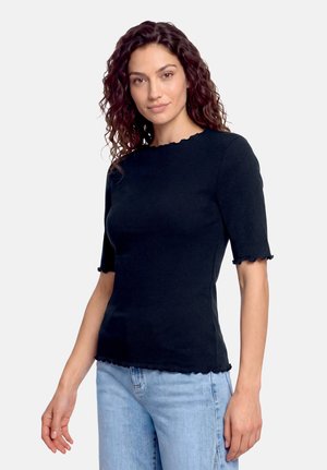Kvinde med krøllet mørkt hår iført en tætsiddende sort kortærmet top med flæsekant og lyseblå jeans, stående mod en ensfarvet baggrund.